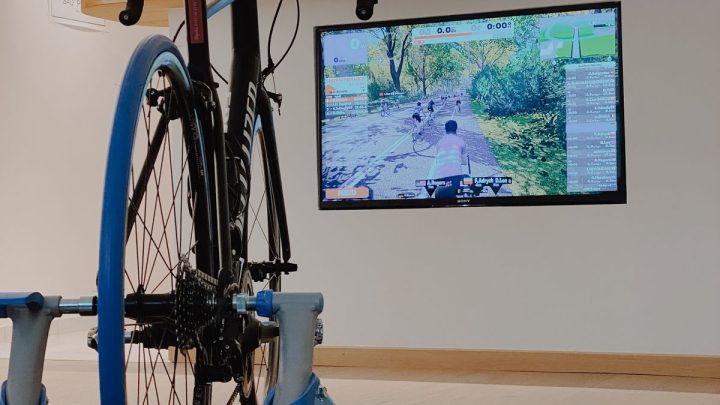 Bicicleta montada en un rodillo inteligente de rueda directa en una sala luminosa, con pantalla mostrando una app de ciclismo virtual