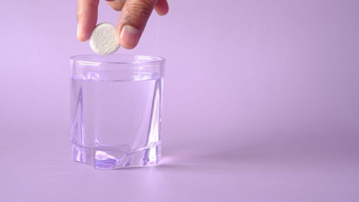 Mano dejando caer una pastilla efervescente en un vaso de agua sobre fondo lila